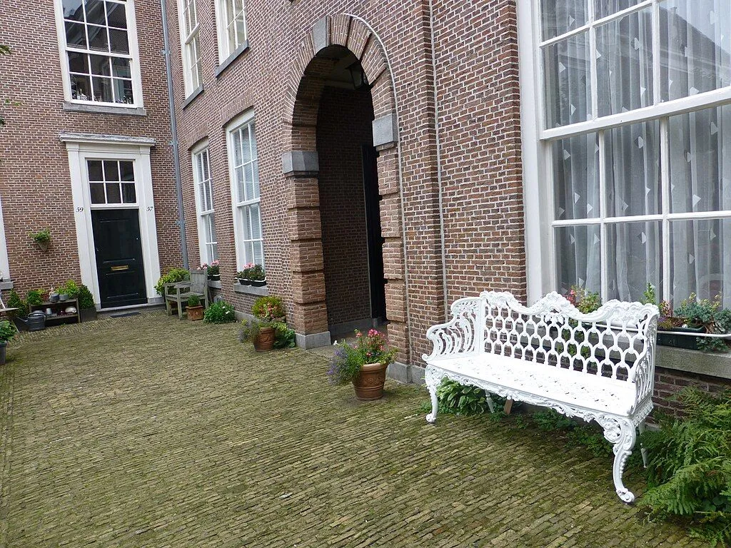 The image captures a charming courtyard scene featuring a traditional red brick building with an arched entryway and an ornate white cast iron bench. The lighting is soft and even, showcasing the textures of the brickwork and the intricate details of the bench. The image is sharp and color-accurate, providing a realistic depiction of the setting. While well-composed with good depth, the perspective could be adjusted for a more dynamic visual impact.