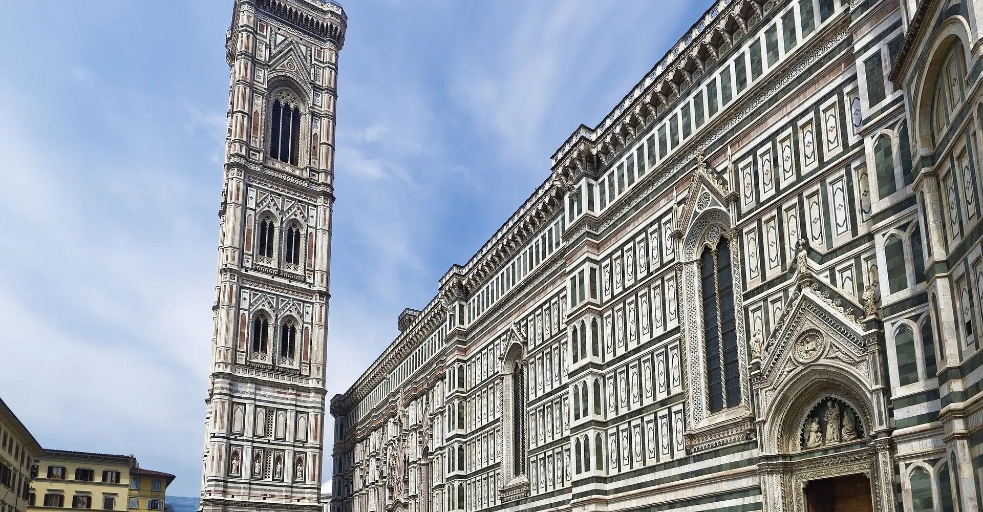 A striking low-angle view of a historic cathedral's highly ornate facade, featuring intricate patterns of white and green marble, alongside its elegant campanile, set against a clear blue sky.