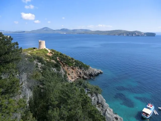 A vibrant and clear image showcasing a historic stone tower perched on a green, rocky hillside overlooking the bright blue sea under a clear sky. Distant landmasses are visible on the horizon, and the water exhibits beautiful variations in blue and turquoise.