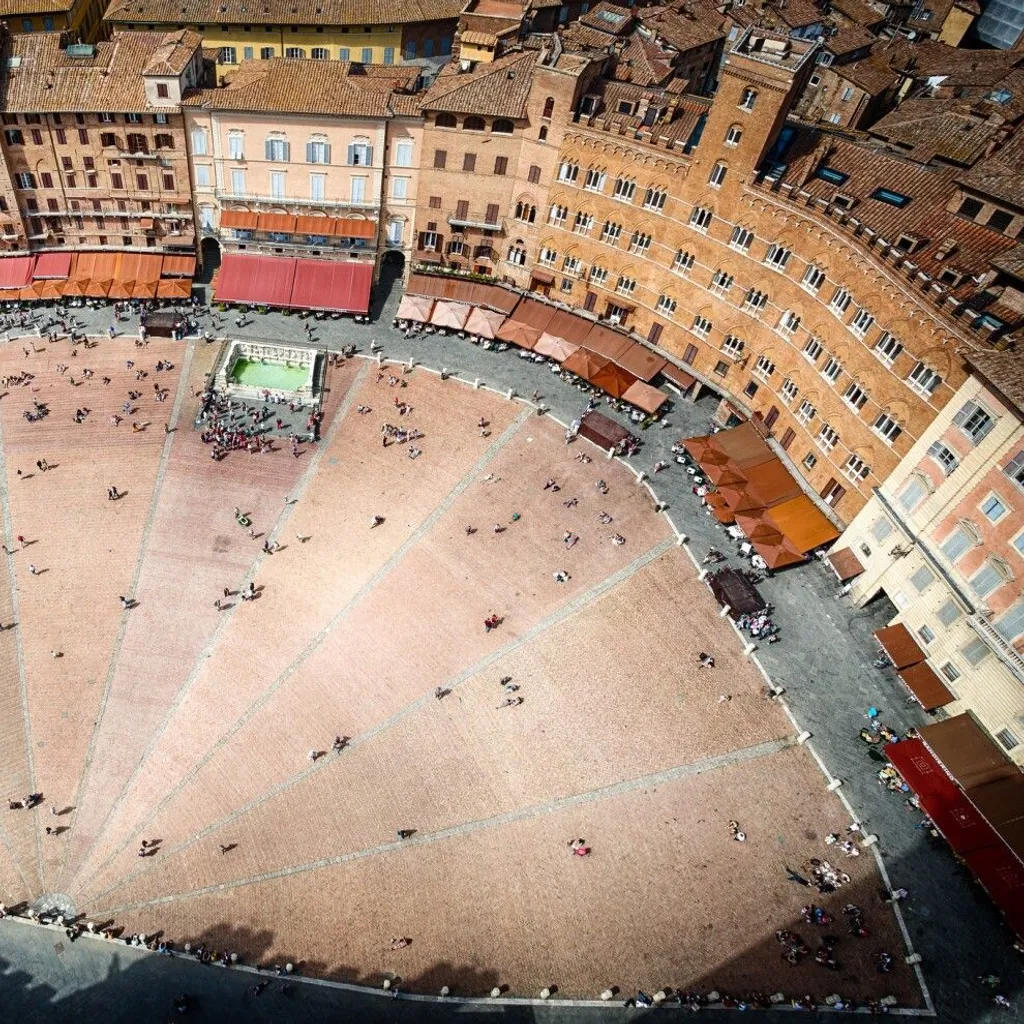 An aerial view captures the unique fan-shaped Piazza del Campo in Siena, Italy, bustling with people. Historic buildings with warm brick and terracotta tones surround the perimeter, with awnings providing shade. The paving pattern of the piazza is clearly visible, leading towards the central fountain area.