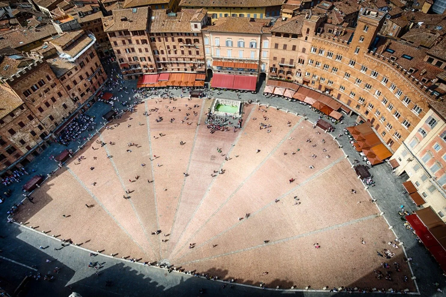 An aerial view captures the unique fan-shaped Piazza del Campo in Siena, Italy, bustling with people. Historic buildings with warm brick and terracotta tones surround the perimeter, with awnings providing shade. The paving pattern of the piazza is clearly visible, leading towards the central fountain area.