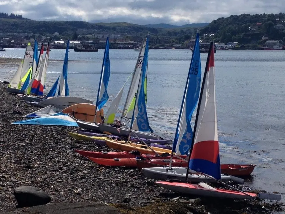 A collection of colorful small sailboats and kayaks resting on a rocky shore next to a body of water, with distant land and buildings visible under a cloudy sky.
