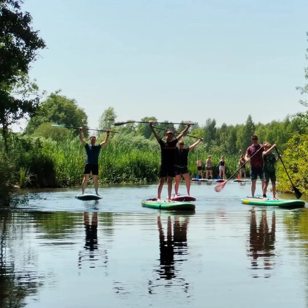 A group of people are enjoying stand-up paddleboarding on a calm river or canal, surrounded by lush green foliage under a clear, sunny sky. Their reflections are visible in the water.