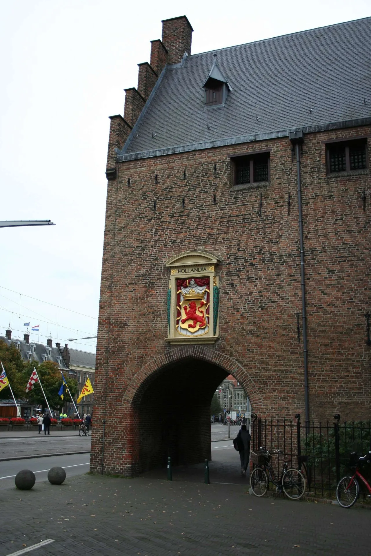 An exterior view of a historic brick building featuring an arched passage and a decorative coat of arms (Hollandia) above it. The scene includes street elements, flags, and people in the background, captured on what appears to be an overcast day.