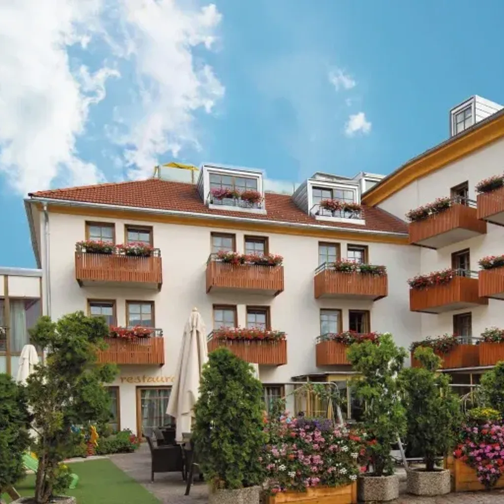 An outdoor view of a multi-story building featuring numerous balconies with flower boxes, surrounded by lush green trees and colorful potted plants. A bright blue sky with white clouds is visible above.