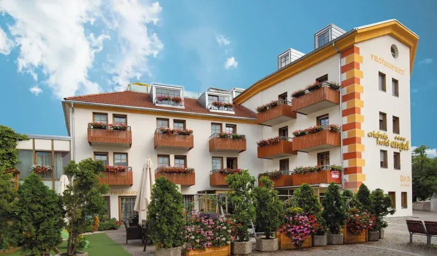 An outdoor view of a multi-story building featuring numerous balconies with flower boxes, surrounded by lush green trees and colorful potted plants. A bright blue sky with white clouds is visible above.