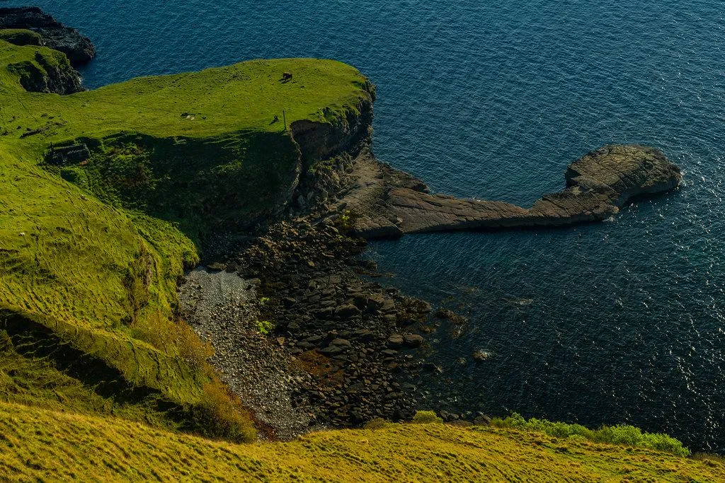 A high-angle view captures a dramatic coastal landscape, showcasing a verdant green cliff dropping to a rocky shore alongside deep blue ocean waters. A natural rock formation extends into the sea, adding to the rugged beauty of the scene.