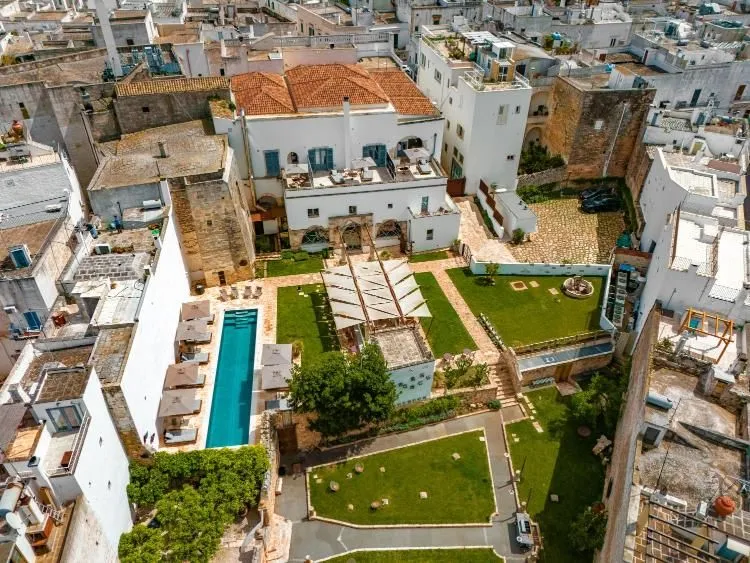 An impressive aerial shot showcasing a large, traditional white-walled property with terracotta roofs, a prominent blue swimming pool, and meticulously maintained green gardens and patios, nestled within a dense urban landscape of similar architecture.
