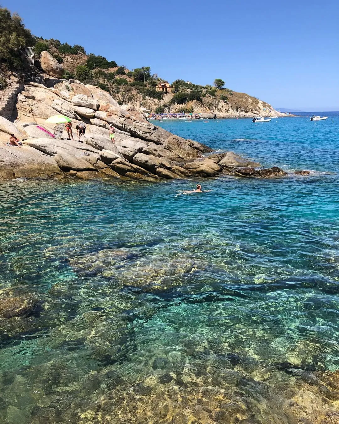 The image displays a clear blue coastal scene with rocky shores and transparent water. The natural lighting highlights the vibrant colors of the sea and the texture of the rocks.