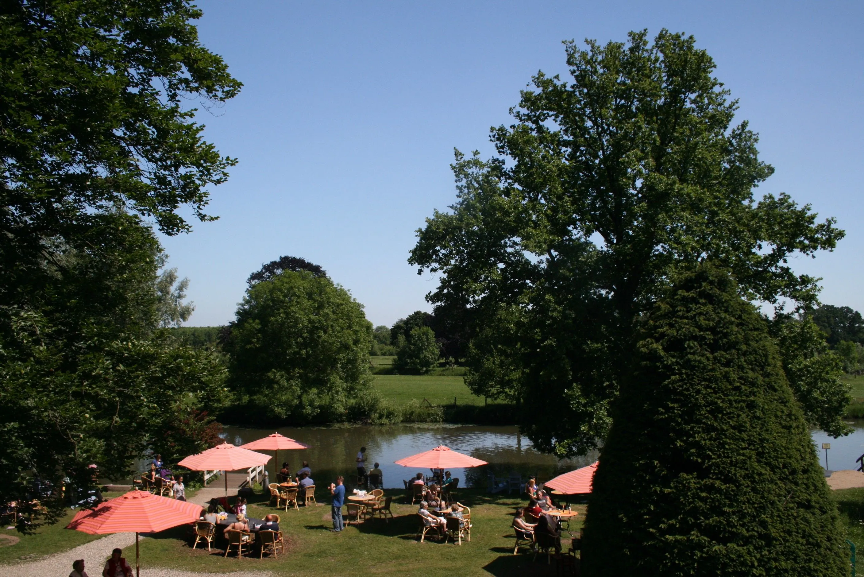 The image captures a beautiful outdoor dining scene by a lake under clear blue skies. The lighting is excellent, providing a natural and vibrant look to the trees, water, and orange umbrellas. The composition is well-balanced, framing the dining area and lake between lush greenery. While the image is photo-realistic and colors are accurate, some finer details, particularly in the distant foliage, appear slightly soft, affecting overall sharpness.