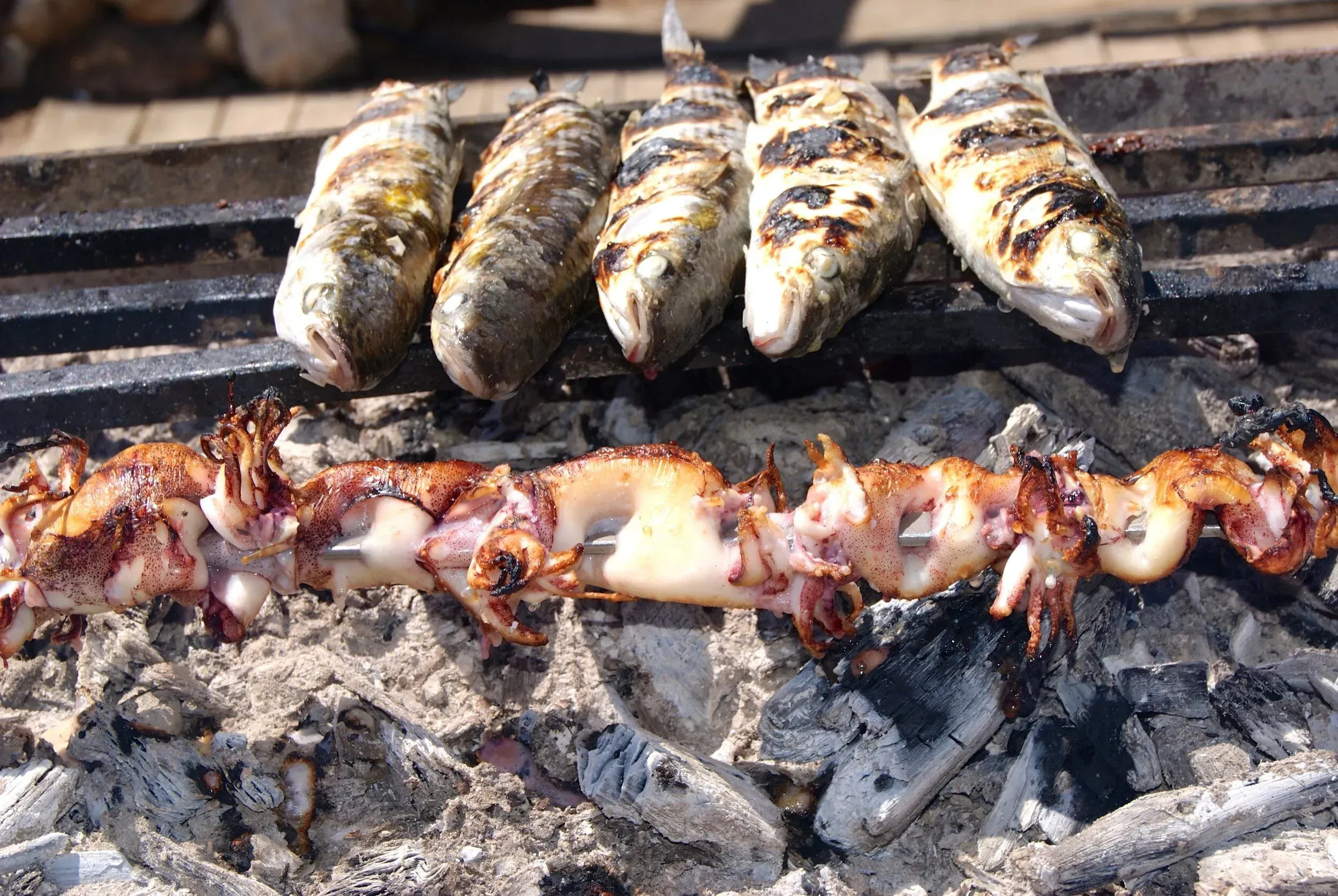 A close-up shot of grilled fish and squid cooking over hot coals. The image clearly captures the texture and color of the cooked seafood.
