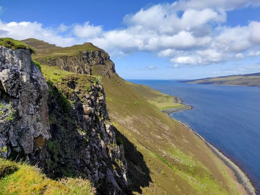 This is a high-quality editorial image of a dramatic coastal landscape featuring cliffs, green slopes, and a calm sea under a partly cloudy sky. The composition is strong, using leading lines and good depth to capture the vastness of the scene.