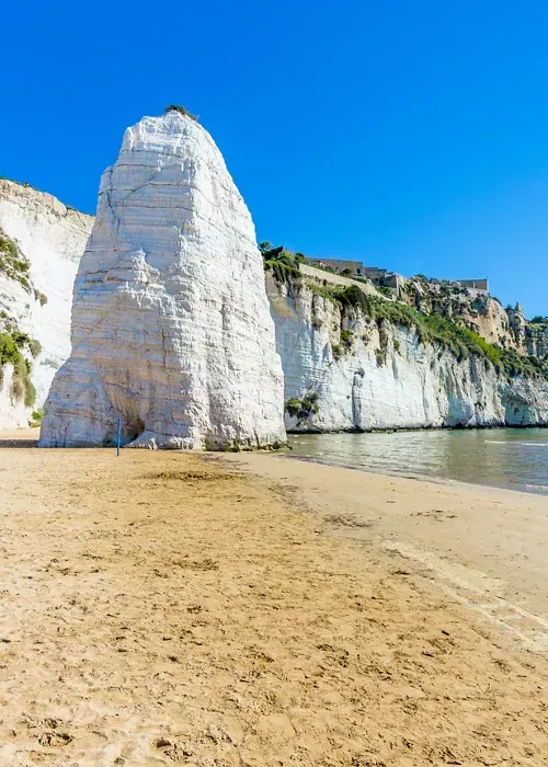 An editorial-style image capturing a stunning coastal landscape featuring a large, prominent white rock formation on a sandy beach under a clear blue sky, with white cliffs extending along the coastline and calm sea waters.