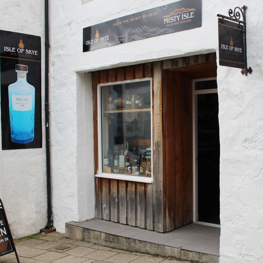 The image shows the exterior of a distillery shop. The composition is acceptable, but could be improved. The lighting is adequate, but not exceptional. The image is reasonably sharp and color-accurate. The key elements, such as the signs and the shop window, are in focus. It is photo-realistic. The image contains promotional signage which is allowed. Well-composed (20/25), Well-lit (12/15), Sharp (16/20), Color-accurate (8/10), Photo-realistic (14/20), Key elements in focus (5/10).