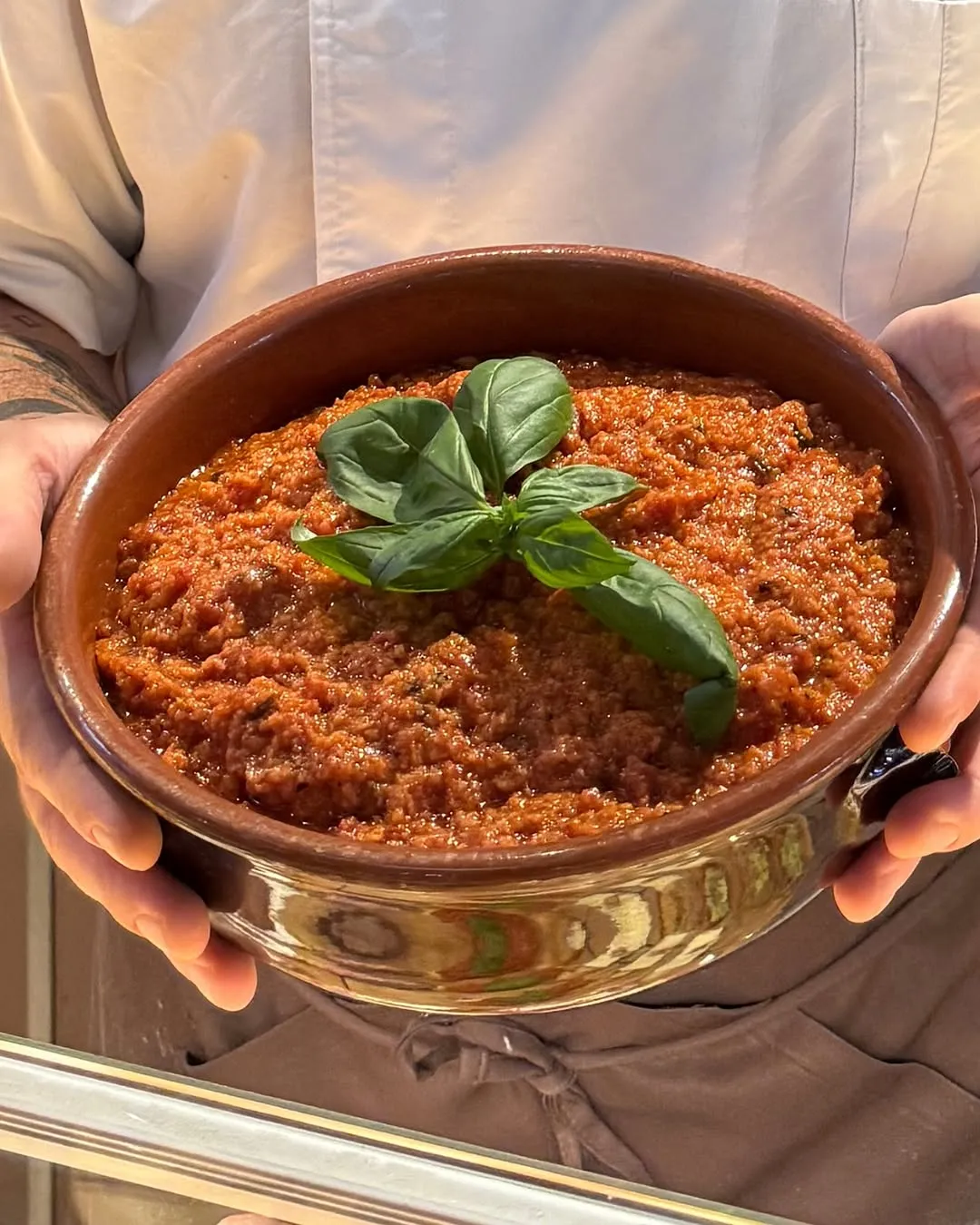 A close-up shot of a rustic bowl filled with a rich red sauce, likely a pesto or similar spread, garnished with fresh green basil leaves. The bowl is held by a person, whose hands are visible, adding a human element to the presentation. The background is softly blurred, keeping the focus on the food.