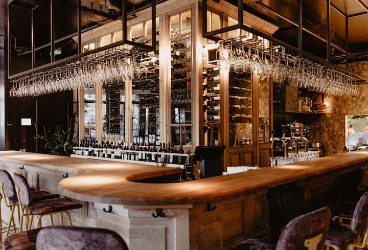 A well-composed and aesthetically pleasing photograph of a sophisticated bar interior. The lighting is warm and even, highlighting the wooden bar counter and extensive wine collection. The image is sharp with accurate colors, and all key elements, such as the hanging glasses and bottled wines, are clearly in focus, contributing to a high degree of photo-realism.