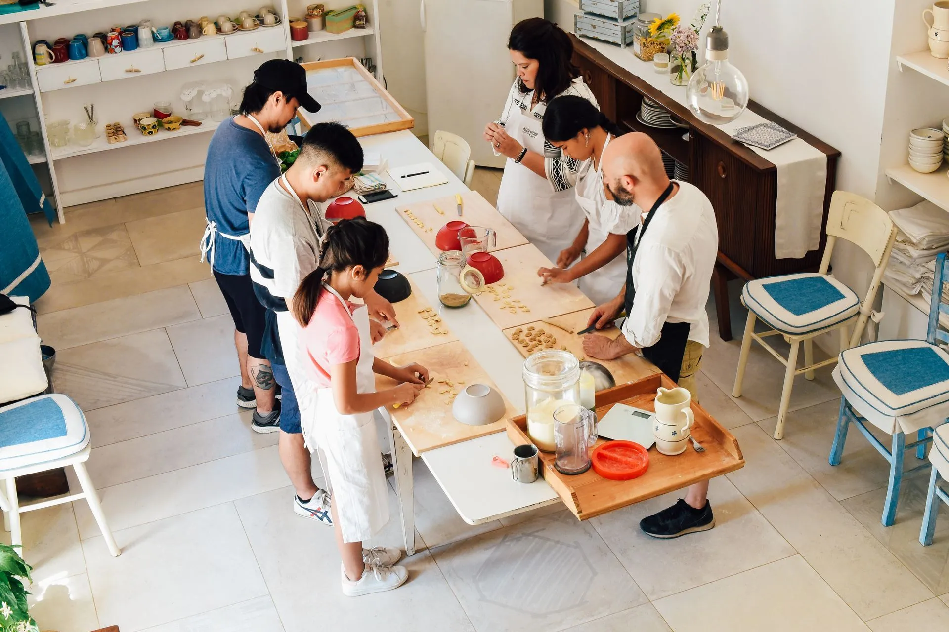 An overhead shot capturing a group of people, including adults and possibly a child, participating in a culinary workshop, likely making pasta or dumplings. They are gathered around a long white table, actively engaged in shaping dough. Various tools and ingredients are spread across the table.