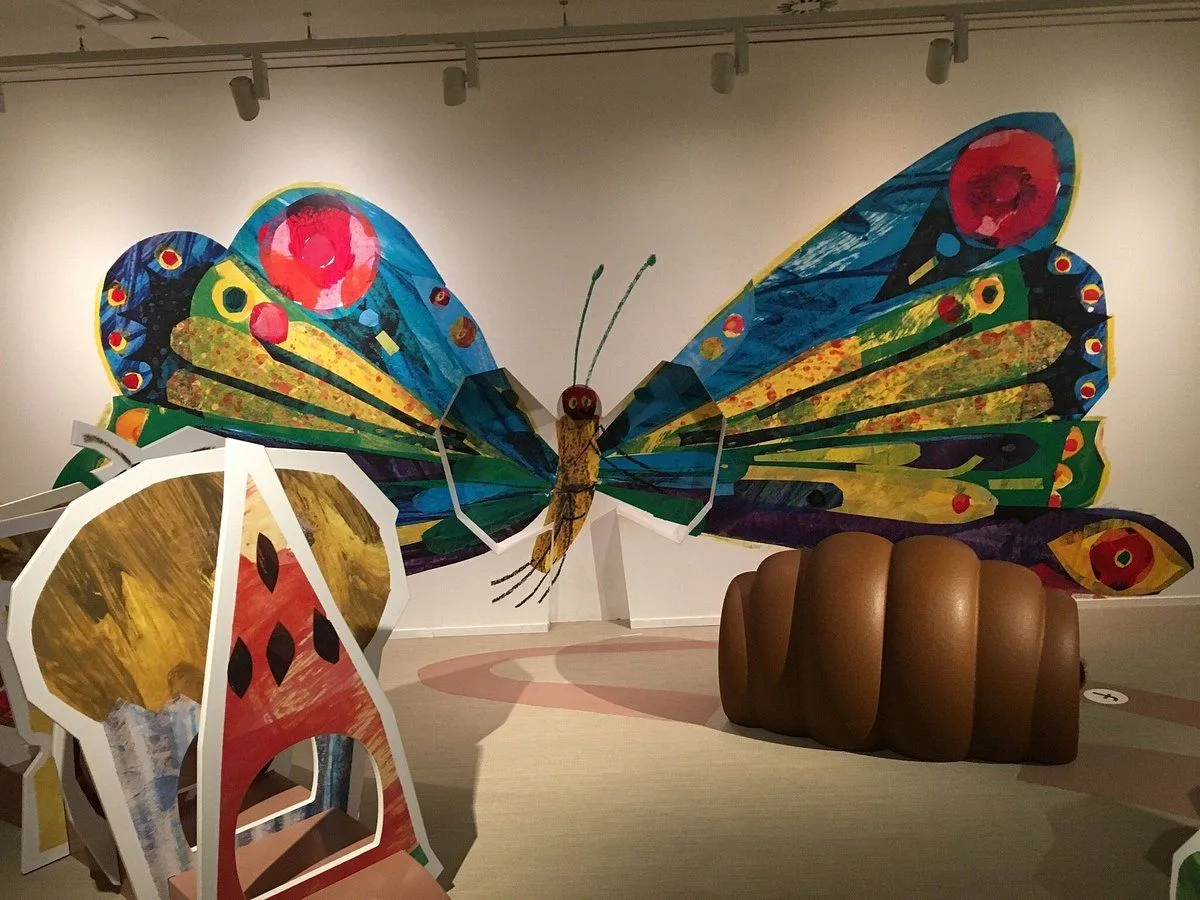 An indoor view of an art installation inspired by 'The Very Hungry Caterpillar', featuring a large, colorful butterfly mural or sculpture on a white wall, accompanied by a large brown caterpillar-like sculpture on the floor and a partially visible, stylized watermelon structure.