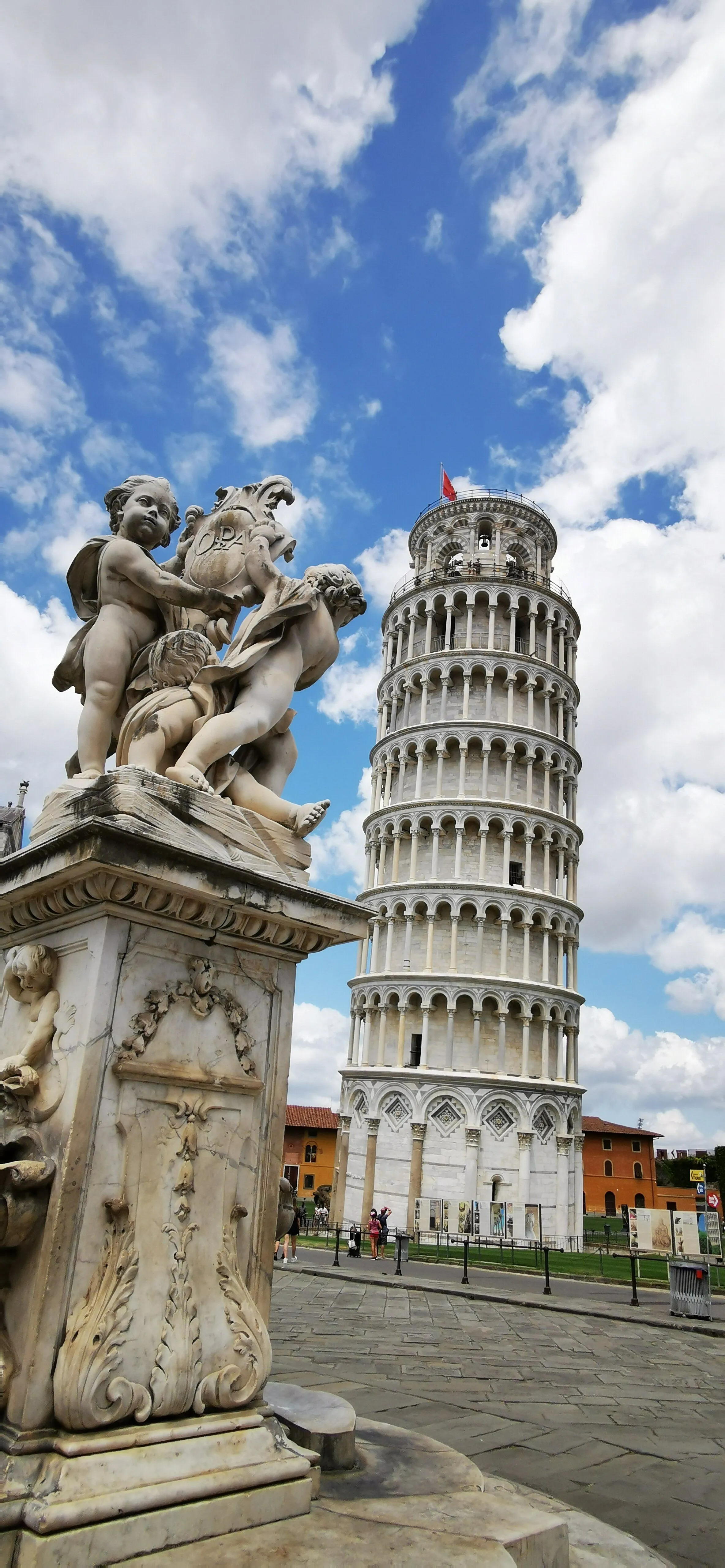 An editorial style image showcasing the Leaning Tower of Pisa in the background, framed by a classical marble sculpture featuring cherubs in the foreground. The sky is blue with scattered white clouds.