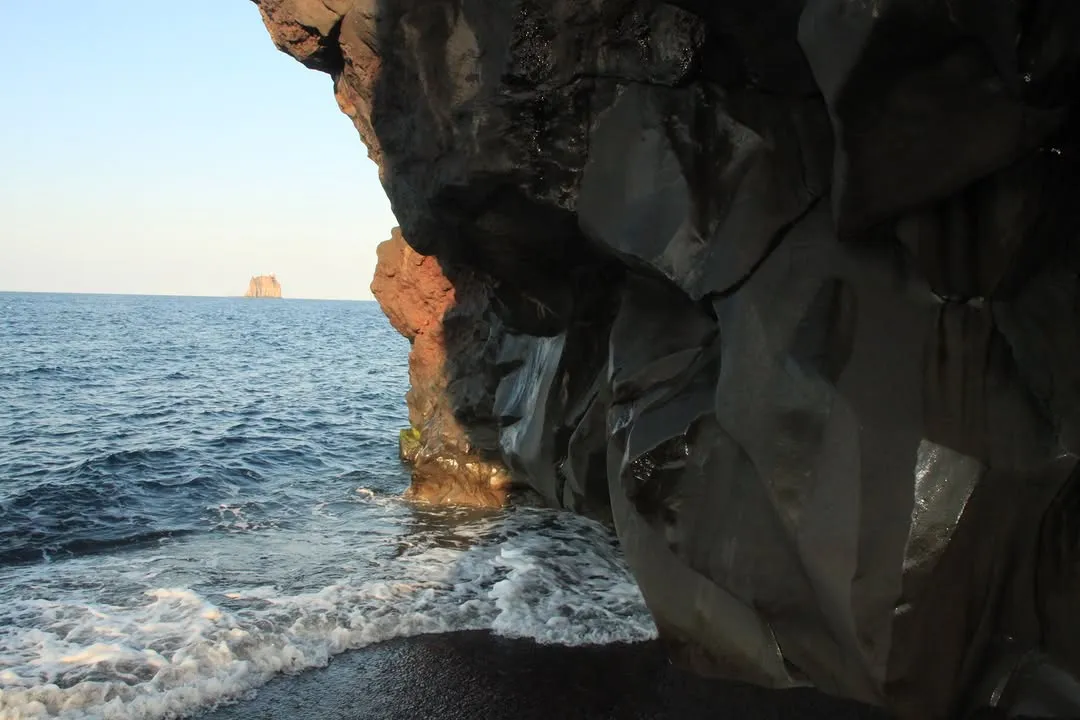 An image capturing a rocky coastline with waves breaking on the shore, viewed from under a dark rock overhang. A distant island is visible on the horizon under a clear sky.