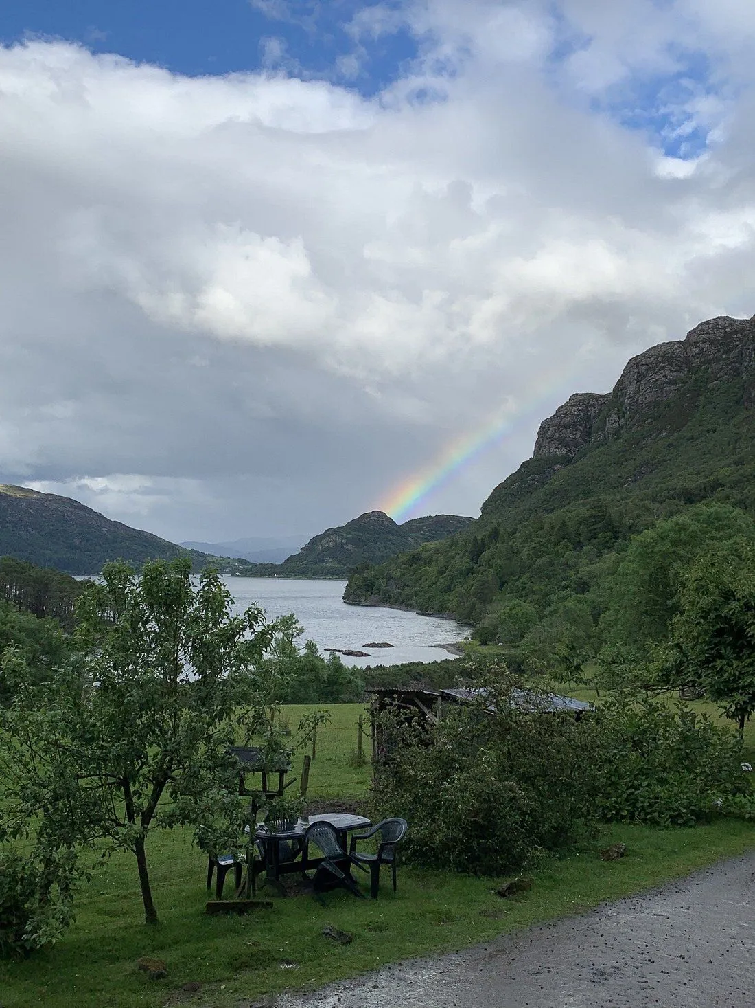 The image captures a beautiful natural landscape featuring a lake, mountains, and a prominent rainbow under an overcast sky. The scene is genuinely photo-realistic and the key elements are in focus.