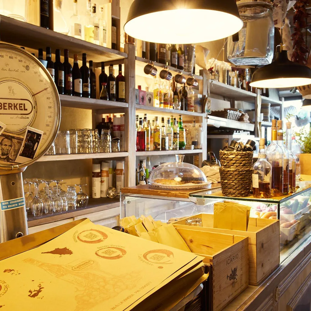 An inviting interior shot of a rustic shop or bar, featuring shelves laden with bottles, glassware, and an antique Berkel scale in the foreground. The warm lighting highlights the various products and the rich texture of the wooden shelves and counter.