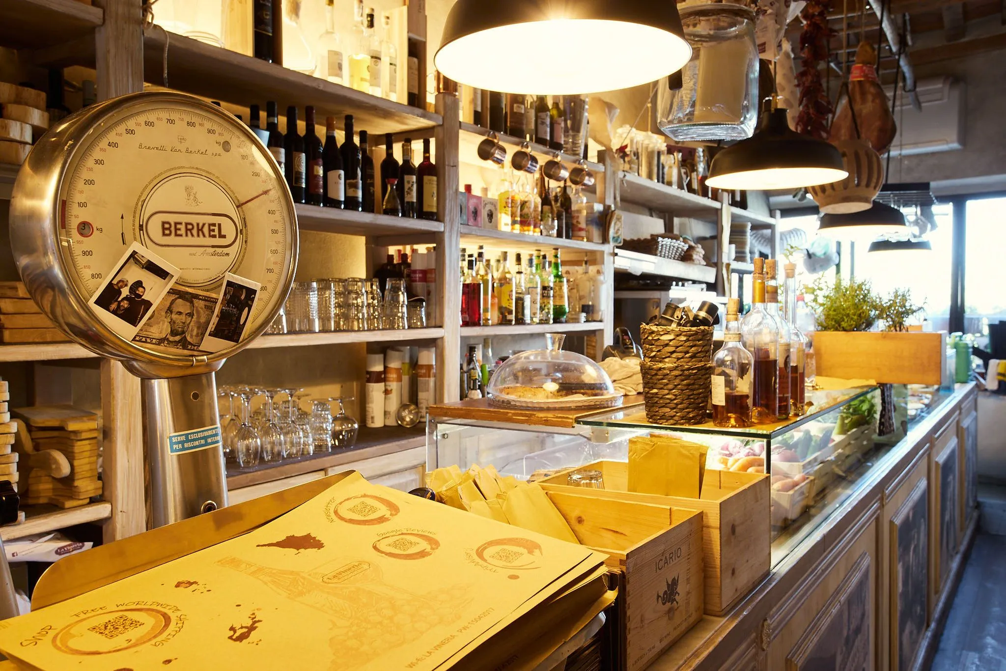 An inviting interior shot of a rustic shop or bar, featuring shelves laden with bottles, glassware, and an antique Berkel scale in the foreground. The warm lighting highlights the various products and the rich texture of the wooden shelves and counter.