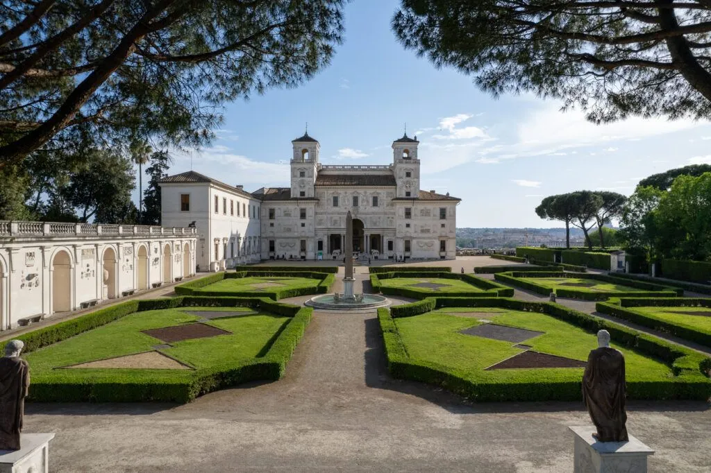 The image is a high-quality, editorial-style photograph of a grand building and its meticulously maintained gardens. It exhibits excellent composition, utilizing natural framing from the trees and leading lines from the garden paths to draw the viewer's eye towards the central architectural subject. The lighting is optimal, providing clear visibility and natural illumination without harsh shadows. The image is sharp with accurate colors, contributing to its strong photo-realistic quality. All key elements, from the intricate details of the building to the manicured hedges, are in sharp focus, making it a visually appealing and well-executed shot.