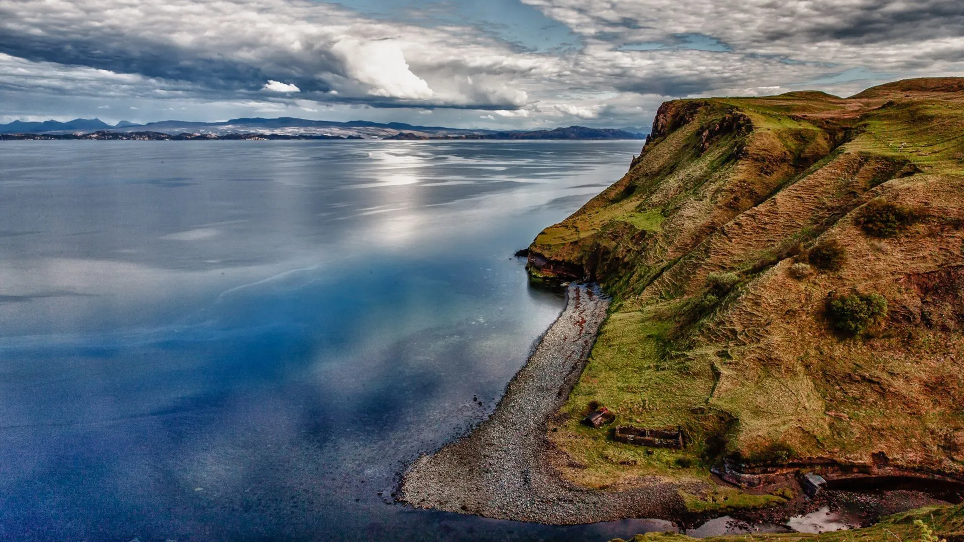 An expansive aerial view of a rugged coastline, featuring a steep, grassy cliff descending into calm, reflective water. A rocky beach curves along the base of the cliff, with distant mountains under a cloudy sky.