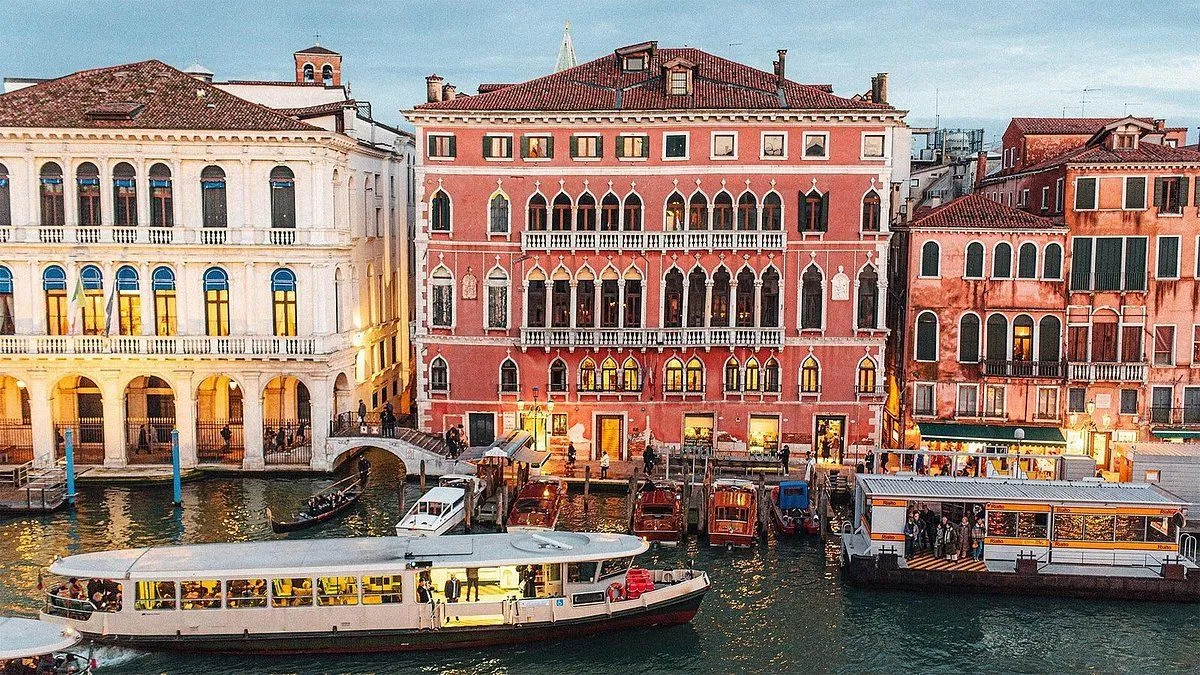 An evocative and well-composed image capturing the vibrant atmosphere of Venice during evening hours. The grand building, illuminated by internal lights, stands prominently alongside the canal bustling with boats and subtle human activity. The overall lighting contributes to a warm, inviting mood.