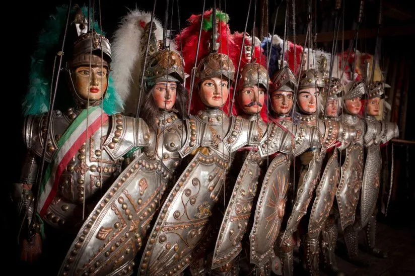 A close-up photograph of several detailed marionettes, dressed in elaborate historical armor, arranged in a row. The lighting highlights the metallic textures and facial features of the puppets.