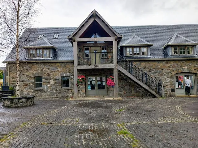 The image features a traditional stone building with a prominent entrance and a slate roof. Hanging flower baskets add a touch of color to the facade. The foreground consists of a cobbled courtyard.