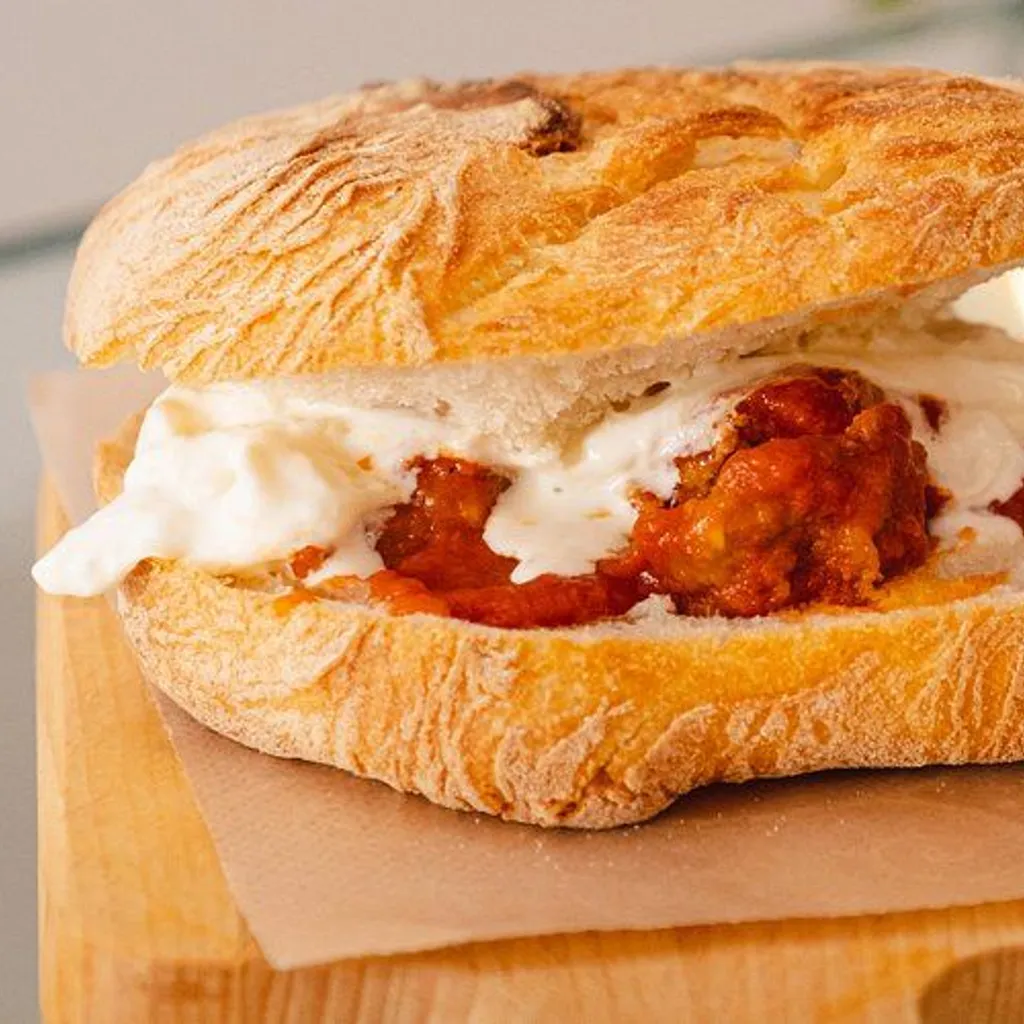 A professionally taken close-up image of a sandwich, likely a focaccia, filled with meatballs or a similar protein in a red sauce, and what appears to be melted white cheese or a creamy sauce. The sandwich is placed on a wooden board with a piece of parchment paper. The background is blurred with bokeh lights.
