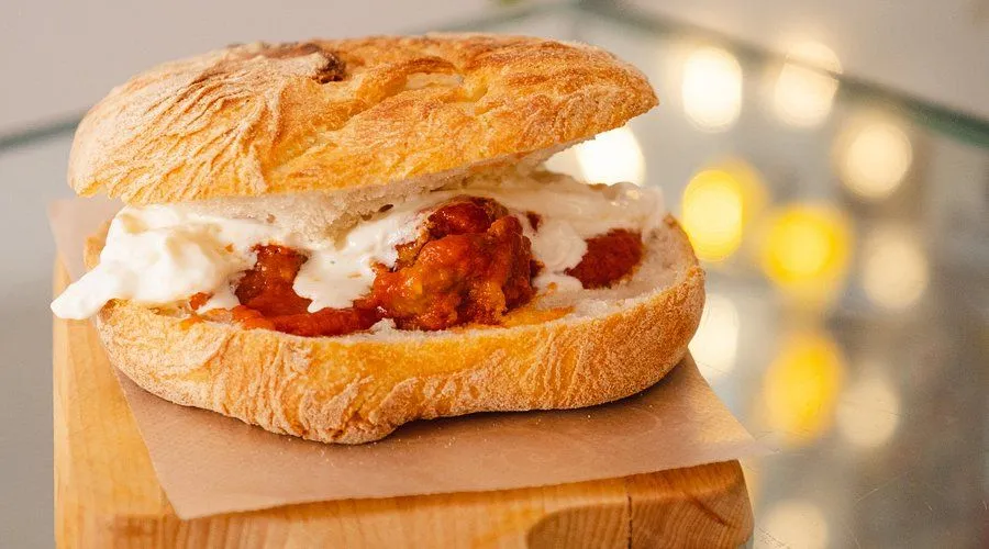 A professionally taken close-up image of a sandwich, likely a focaccia, filled with meatballs or a similar protein in a red sauce, and what appears to be melted white cheese or a creamy sauce. The sandwich is placed on a wooden board with a piece of parchment paper. The background is blurred with bokeh lights.