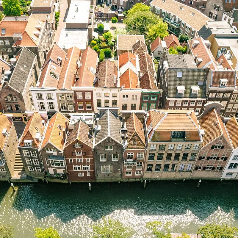 An aerial view capturing a dense row of traditional European houses, likely alongside a canal or river, featuring diverse architectural styles, varying roof colors, and details of windows and facades under bright daylight.