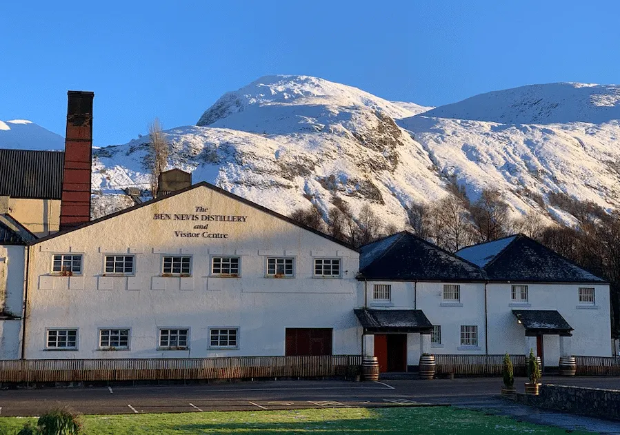 A pristine image of The Ben Nevis Distillery and Visitor Centre, featuring a white building with dark roofs nestled at the base of a majestic, snow-capped mountain under a clear blue sky. The foreground shows a partial parking area and green grass.