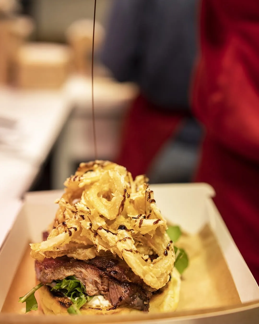 The image features a close-up, professionally taken shot of a food item, likely a burger or steak, topped with crispy onion rings and a sauce being drizzled over it. The food is presented in a takeout container, with a blurred background showing a person in a red apron, suggesting a kitchen or food service environment. The focus is sharp on the food, capturing texture and detail.