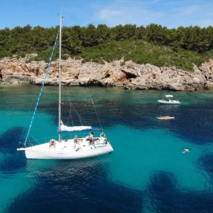 The image captures a beautiful Mediterranean scene with a sailboat anchored in clear, turquoise water near a rocky, tree-lined coast. The vibrant blue hues of the sea transition from light to deep, showcasing excellent clarity. Sunlight bathes the scene, highlighting the details of the boat and the natural environment.