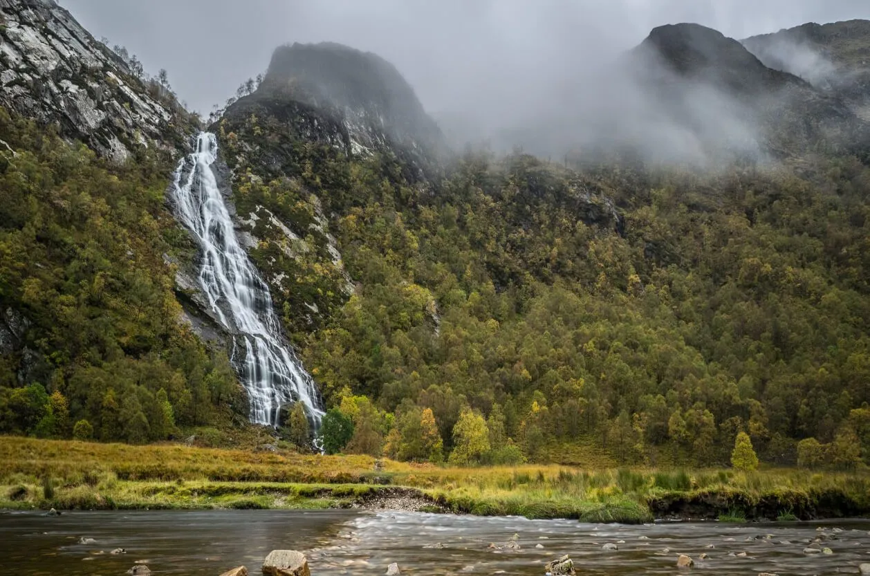 A majestic waterfall cascades down a verdant, tree-covered mountain slope, with a calm river flowing in the foreground under an overcast sky.