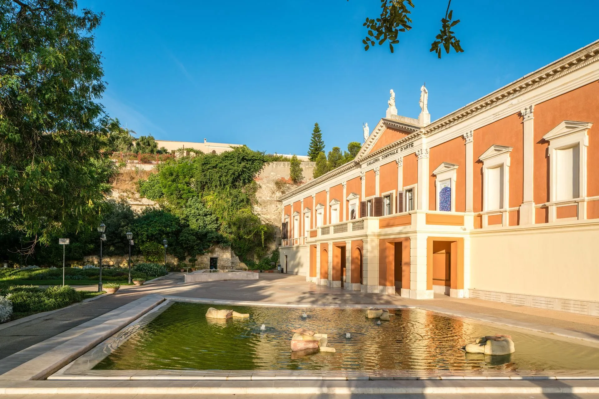 An outdoor shot of a historic building with classical architectural elements, featuring a large courtyard and a decorative fountain in the foreground. The image is well-composed, brightly lit by natural sunlight, and exhibits strong photorealism. The colors are vibrant and accurate, and the key elements, including the building and fountain, are sharp and in focus. The blue sky provides a clear backdrop.