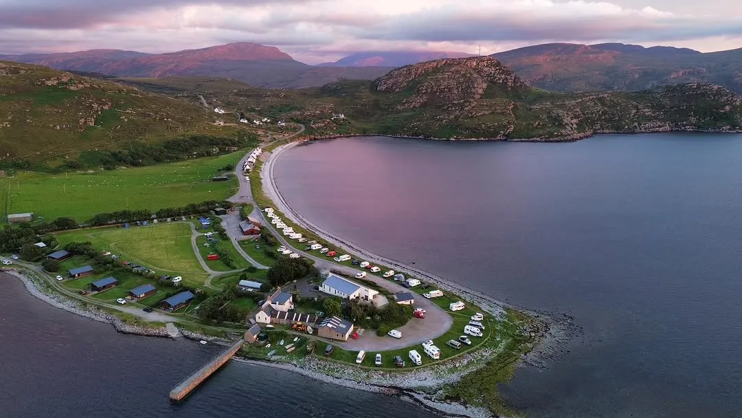 An aerial photograph capturing a picturesque coastal bay with a small village or caravan park nestled along its curved shore, surrounded by rolling hills and mountains under a dramatic sky.