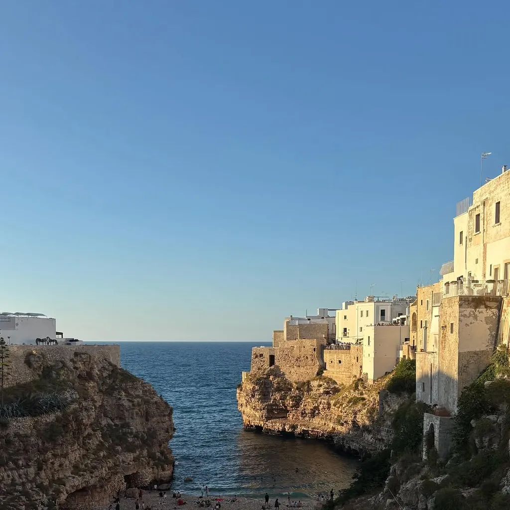 An outdoor, eye-level, wide shot of a scenic Italian coastal town built into cliffs overlooking a popular beach cove during what appears to be golden hour. The clear blue sky meets the horizon of the calm sea. People are visible on the sandy beach below the rugged cliffs.