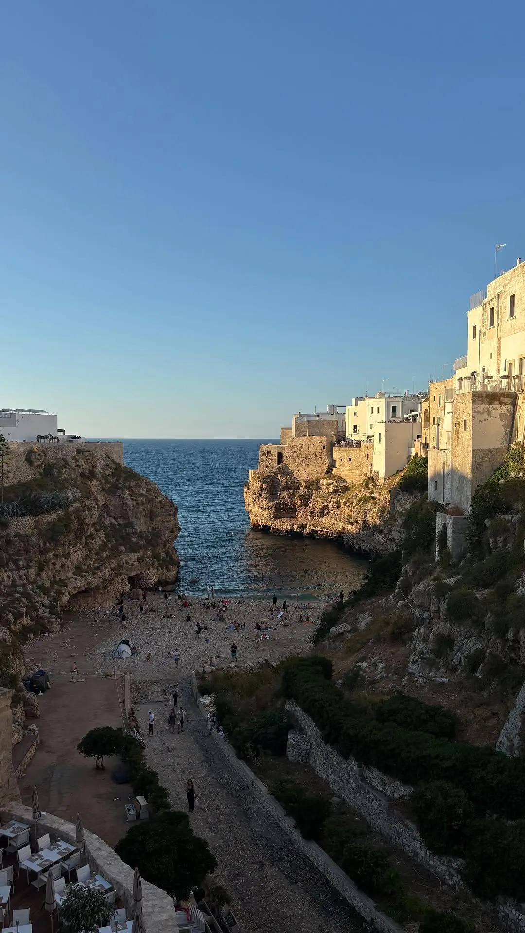 An outdoor, eye-level, wide shot of a scenic Italian coastal town built into cliffs overlooking a popular beach cove during what appears to be golden hour. The clear blue sky meets the horizon of the calm sea. People are visible on the sandy beach below the rugged cliffs.