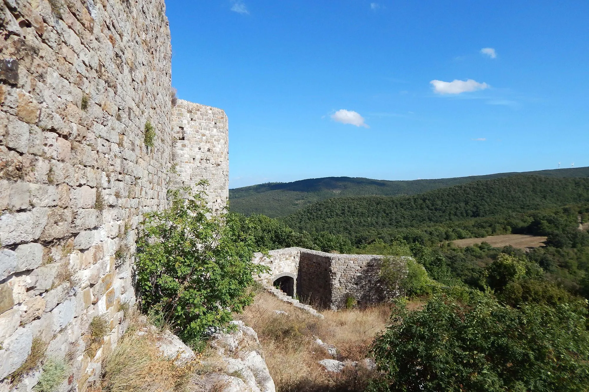 An outdoor shot of ancient stone castle ruins, featuring a tall stone wall on the left, leading to smaller ruined structures and an archway. In the background, green hills and mountains rise under a clear blue sky with sparse clouds. The foreground shows dry grass and scattered green bushes.