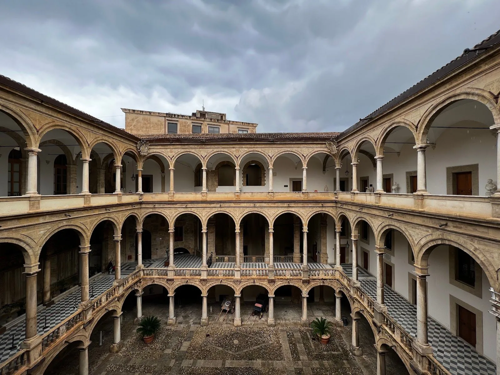 An elevated view of a historic, multi-level courtyard featuring arched colonnades, checkerboard tile pathways on the upper levels, and a central cobblestone area, all under an overcast sky.