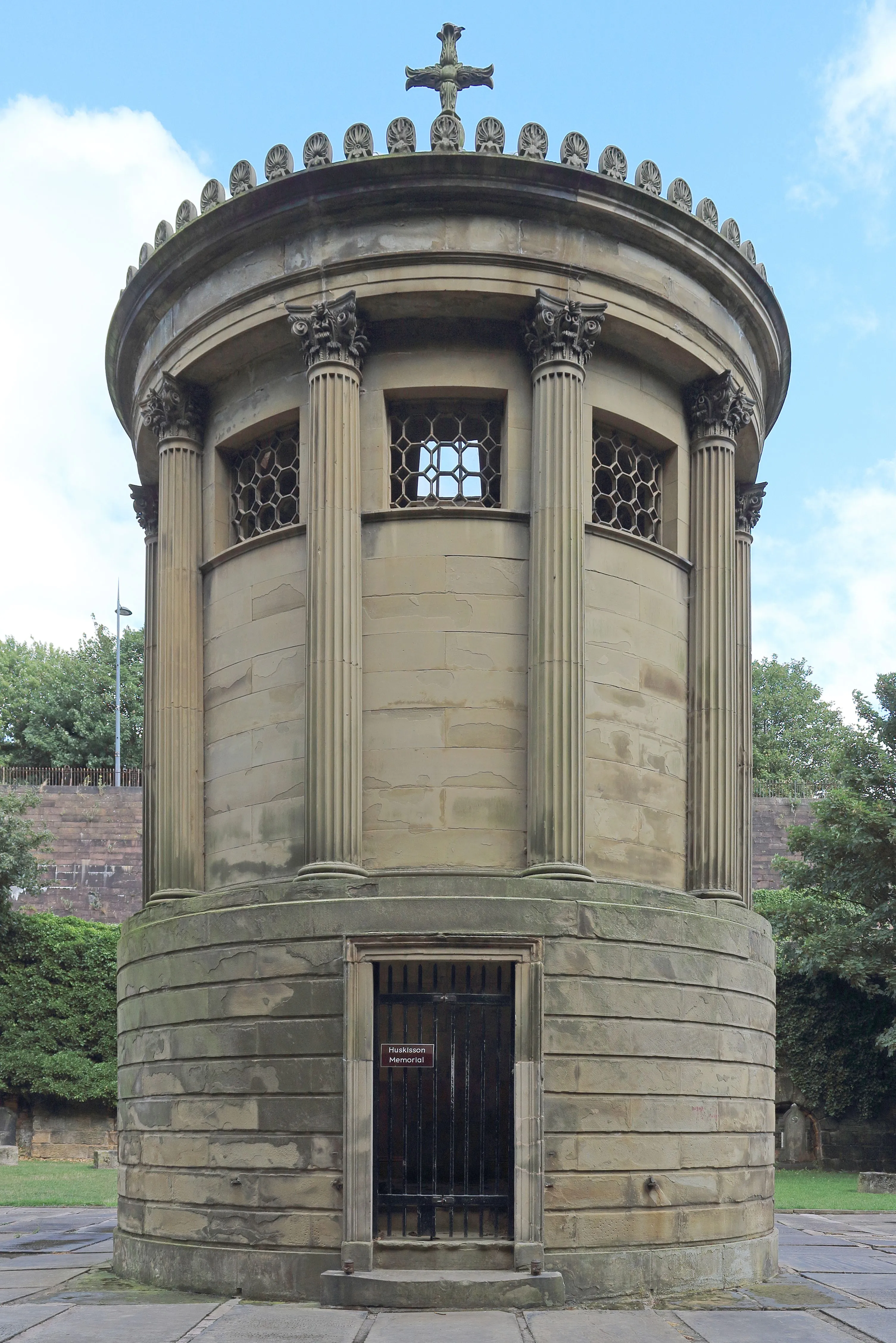 A well-composed and sharp photograph of a circular stone building, possibly a monument or memorial, featuring classical columns, ornate window grates, and a wooden door. The building is made of light-colored stone and is set against a partially visible blue sky and green foliage.