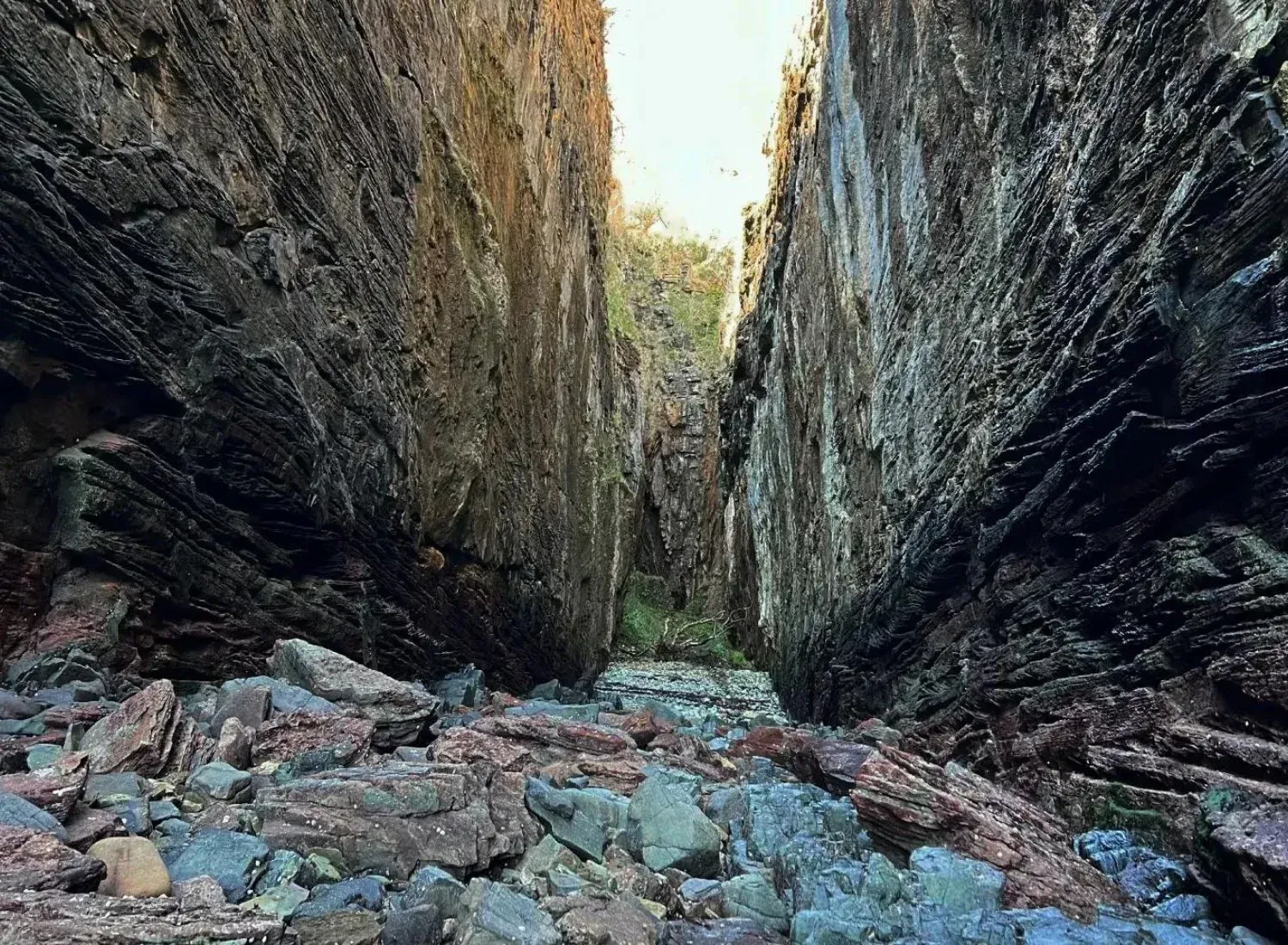A striking image of a narrow rock chasm or gorge, featuring tall, textured rock walls and a foreground covered in various colored boulders. The bright light at the top suggests an open sky, creating a strong sense of depth.