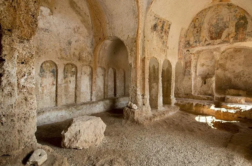 An interior shot of an ancient, possibly rock-cut or cave, structure featuring rough stone walls, arched niches, and some faded frescoes or paintings on the upper walls and ceiling. A large stone rests on the gravelly floor in the foreground, with pillars and further arches visible deeper within the space.