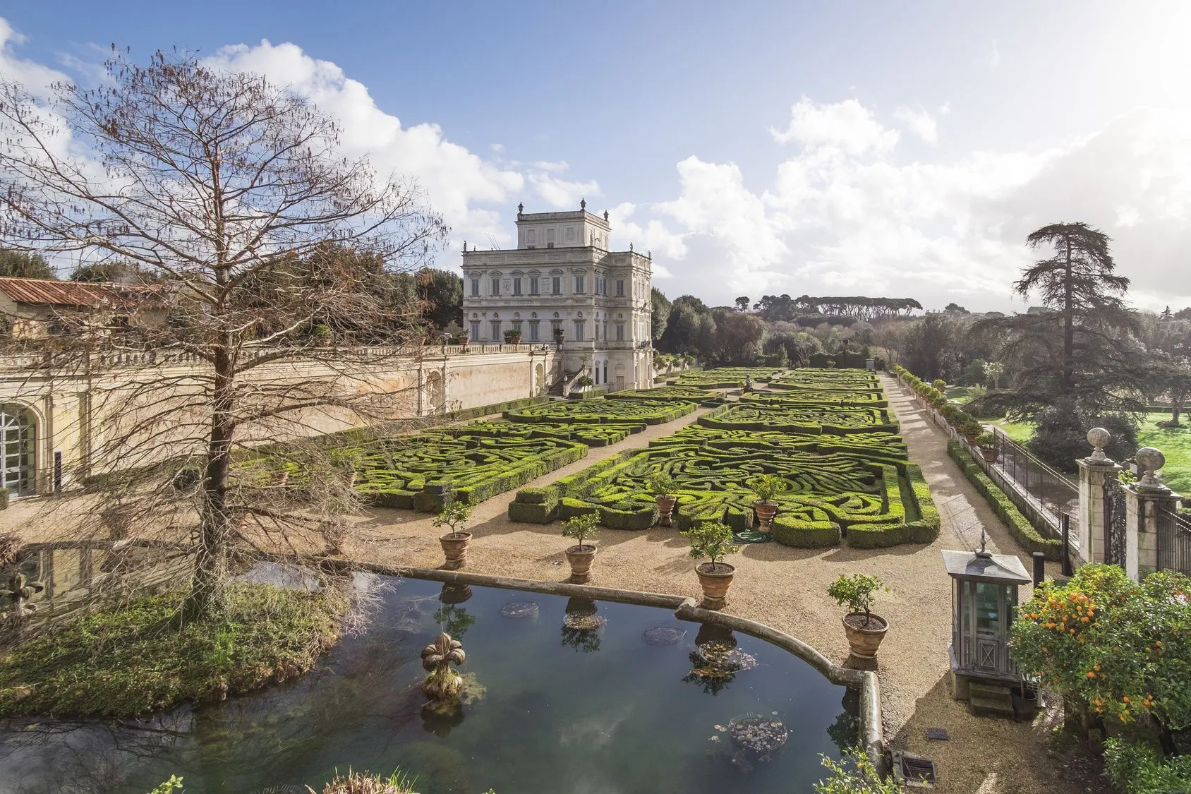 An aerial view capturing the grandeur of Villa Doria Pamphili, featuring its historic building, meticulously designed formal gardens with an intricate hedge maze, and a serene reflecting pool in the foreground. The composition highlights the vastness and detailed landscaping of the estate under a clear sky.