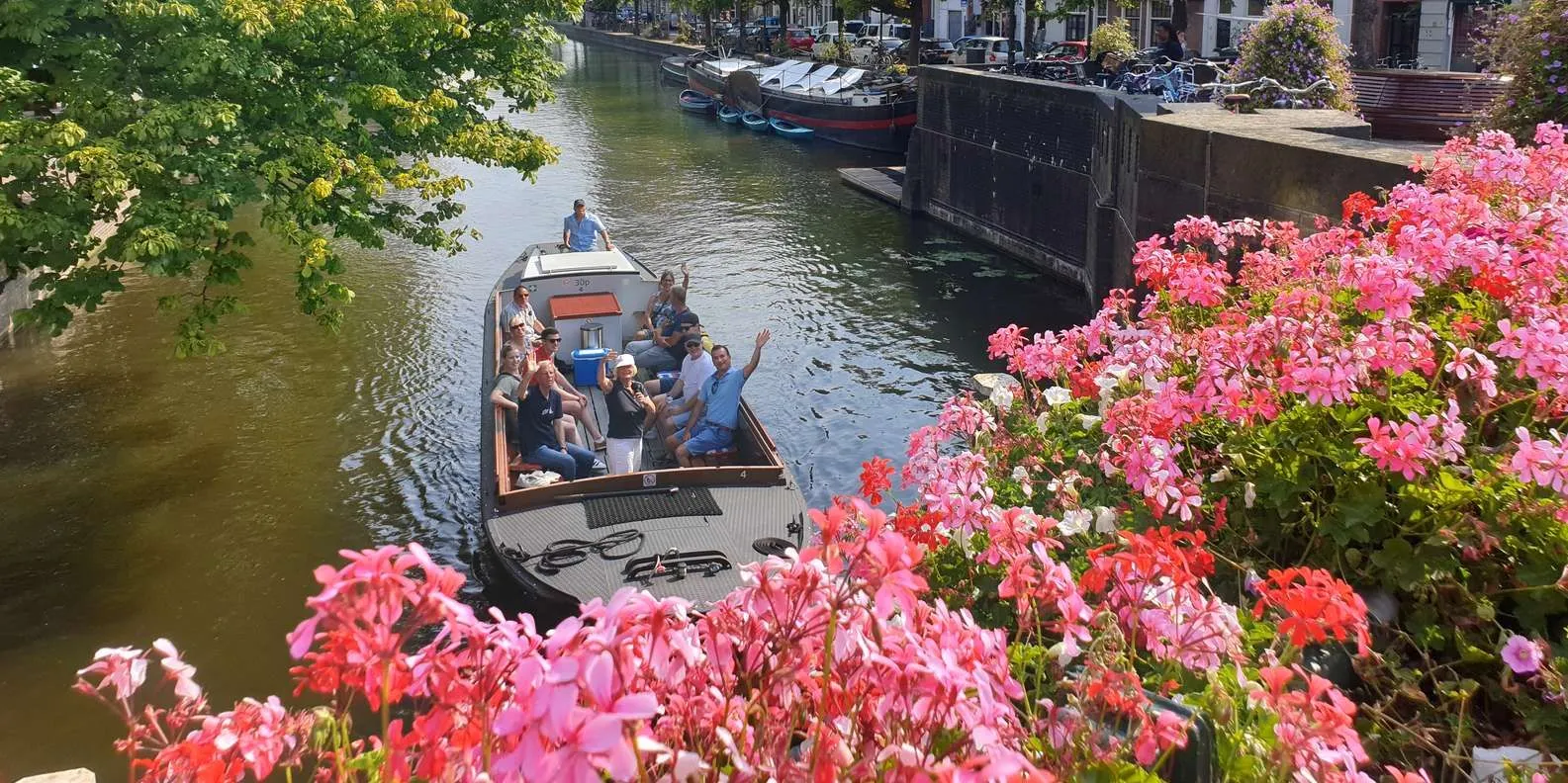The image is well-composed with vibrant foreground flowers providing an attractive frame for the boat on the canal. Natural sunlight illuminates the scene effectively, resulting in good overall lighting and accurate, lively colors. The key elements, including the people on the boat and the framing flowers, are sharp and in clear focus, contributing to a strong photo-realistic aesthetic. The background elements are also reasonably clear, maintaining good depth of field.